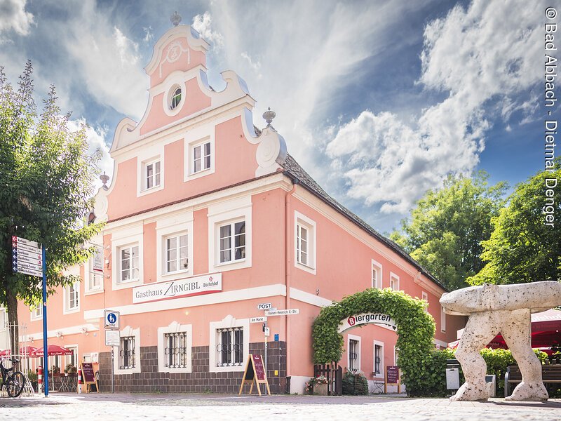 Ein rosa Geb&auml;ude mit Barockfassade, daneben ein Biergarten mit gr&uuml;ner Laube, im Vordergrund eine Steinfigur.