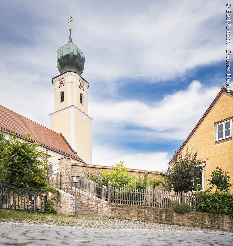 Kirchturm mit gr&uuml;nem Zwiebeldach neben gelbem Geb&auml;ude, umgeben von Garten und Zaun, unter blauem Himmel.