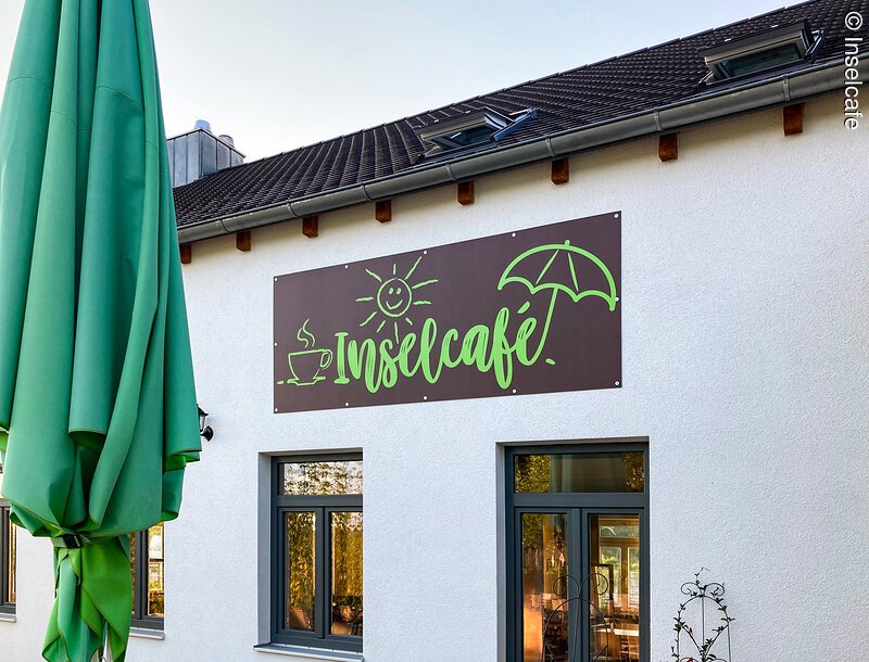 Au&szlig;enansicht eines Geb&auml;udes mit Schild &bdquo;Inselcaf&eacute;&ldquo; und geschlossenem gr&uuml;nem Sonnenschirm davor.