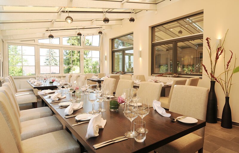 Hell eingerichteter Speiseraum mit gedeckten Tischen und gro&szlig;en Fenstern zu einem Garten hin.