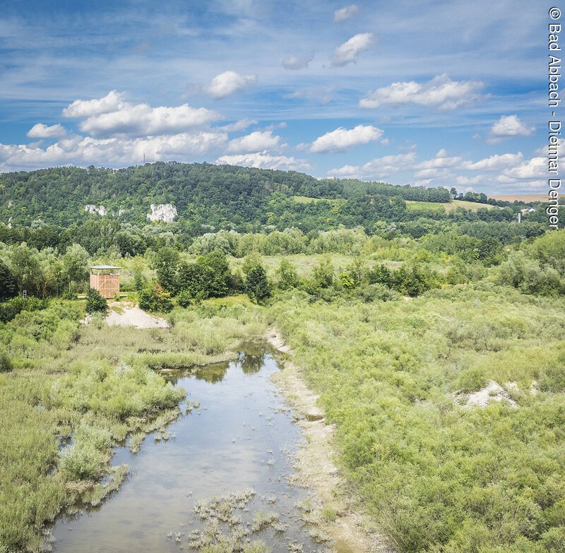 Landschaft mit Fluss, umgeben von B&auml;umen und H&uuml;geln unter blauem Himmel mit Wolken.