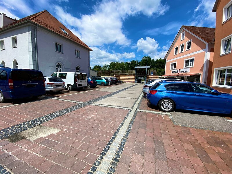Parkplatz mit mehreren geparkten Autos vor zwei Geb&auml;uden unter blauem Himmel mit Wolken