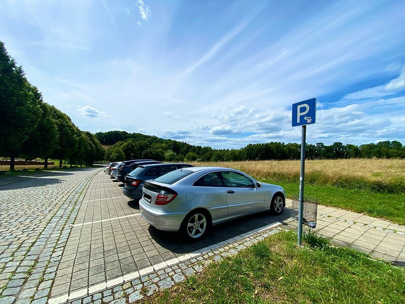 Parkplatz mit mehreren parkenden Autos neben einem Feld und B&auml;umen unter blauem Himmel mit Wolken