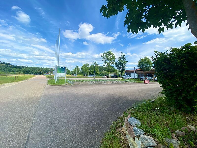 Parkplatz mit Autos und Fahrradst&auml;ndern bei blauem Himmel, links Wiese und Weg, rechts B&auml;ume und Str&auml;ucher