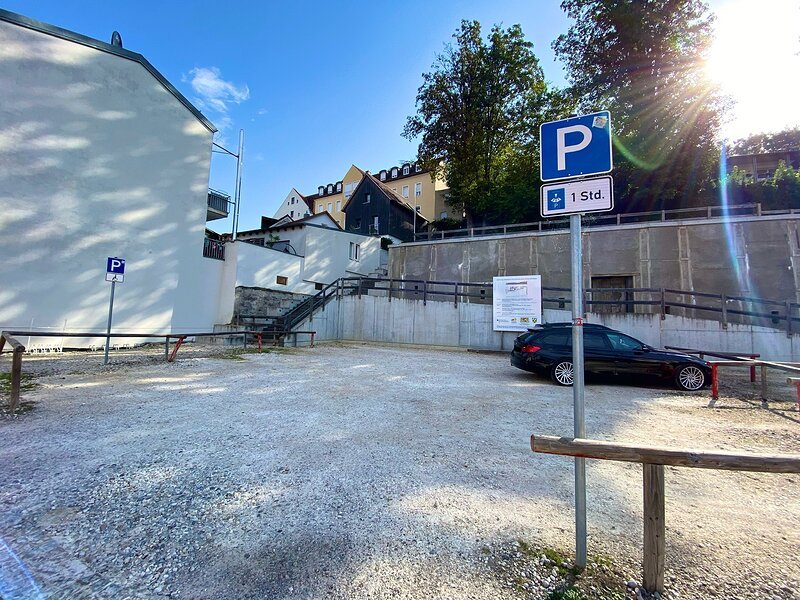 Parkplatz mit Schotterboden, zwei Parkschildern und einem schwarzen Auto, umgeben von Geb&auml;uden und B&auml;umen