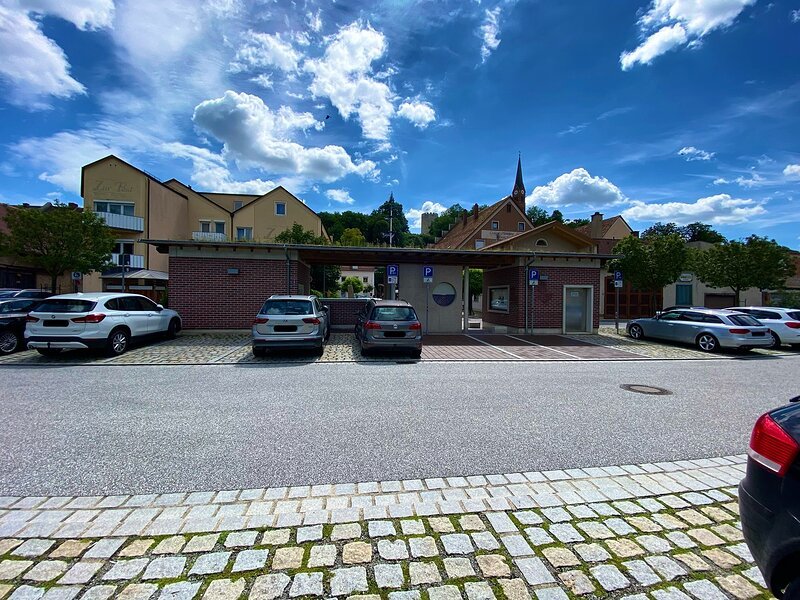 Parkplatz mit mehreren Autos vor einem kleinen Backsteingeb&auml;ude und H&auml;usern unter blauem Himmel mit Wolken