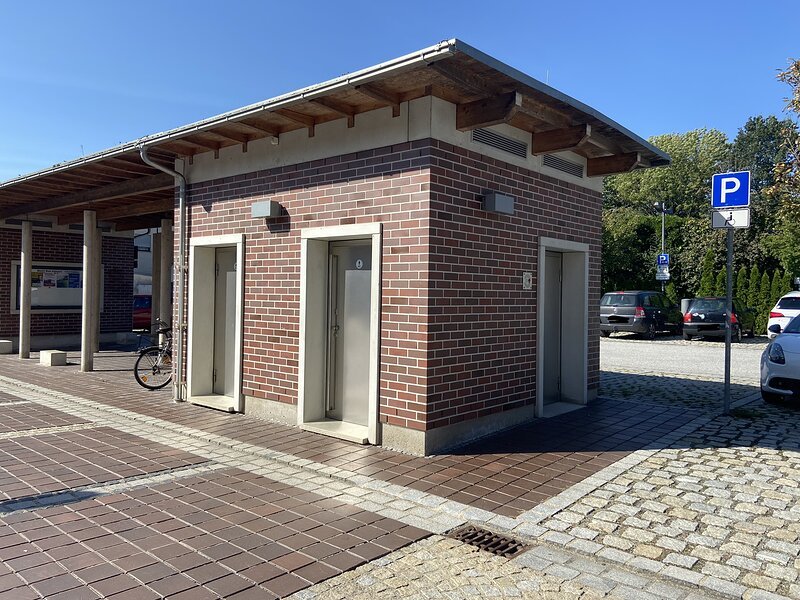 Geb&auml;ude mit drei T&uuml;ren, Parkplatz mit Behindertenparkplatzschild und mehreren Autos im Hintergrund.