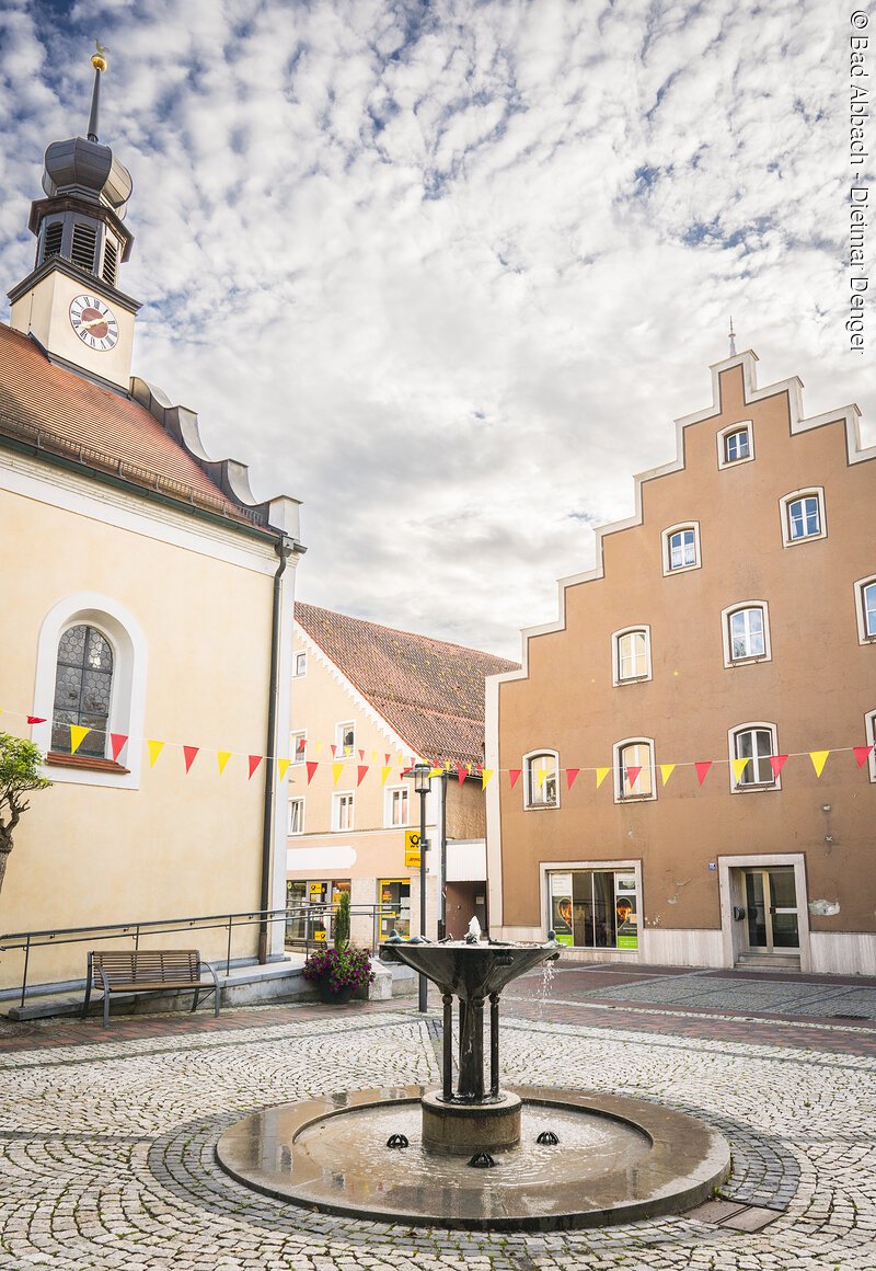 Kleiner Platz mit Brunnen, umgeben von historischen Geb&auml;uden, mit bunten Wimpeln geschm&uuml;ckt, unter bew&ouml;lktem Himmel.