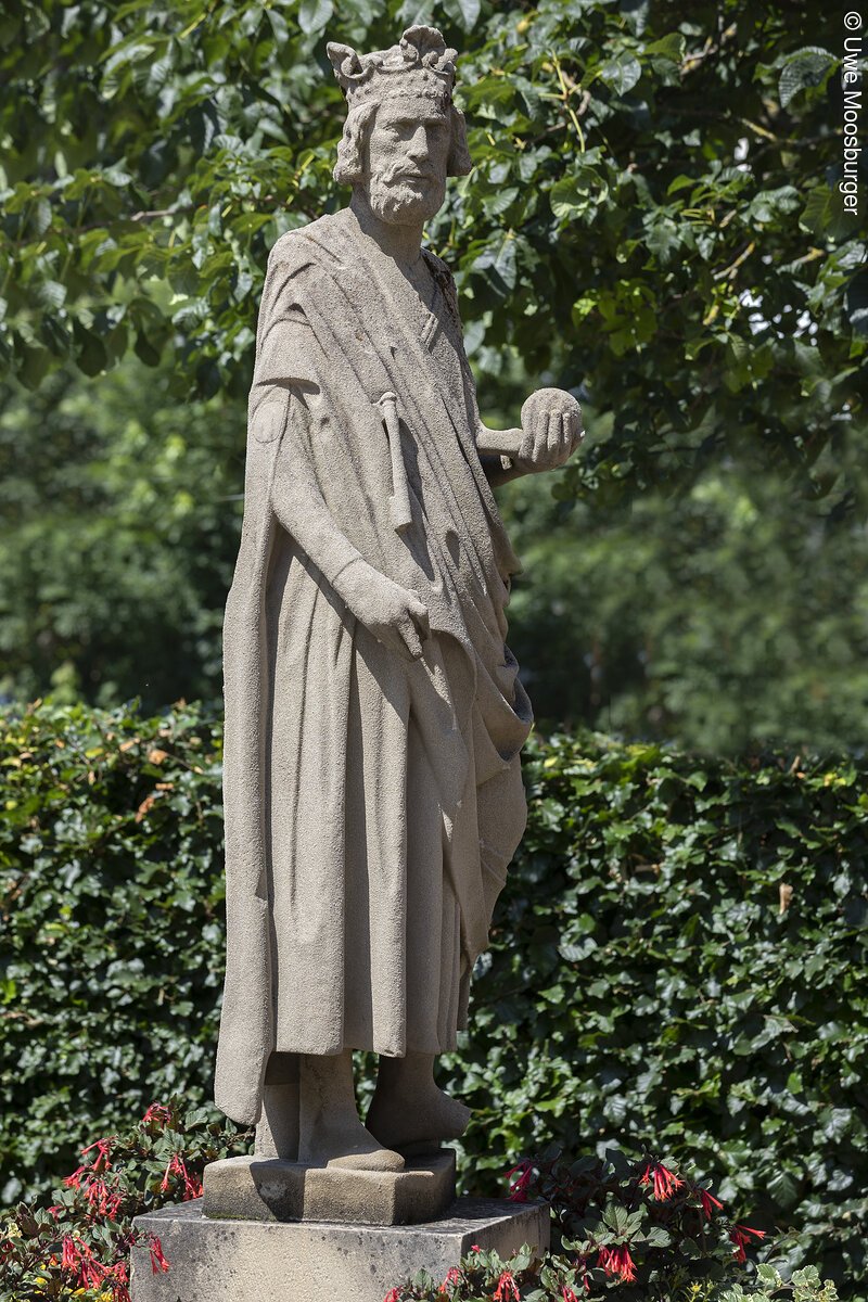 Steinerne Statue eines b&auml;rtigen K&ouml;nigs mit Krone und Kugel in der rechten Hand umgeben von gr&uuml;nen Pflanzen.
