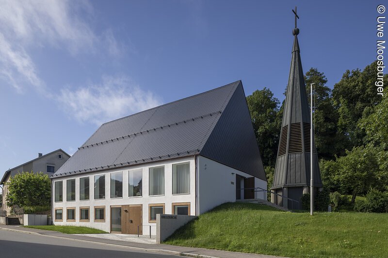 Kirchengeb&auml;ude mit spitzem Dach und separatem Glockenturm mit Kreuz umgeben von B&auml;umen und Rasenfl&auml;che.