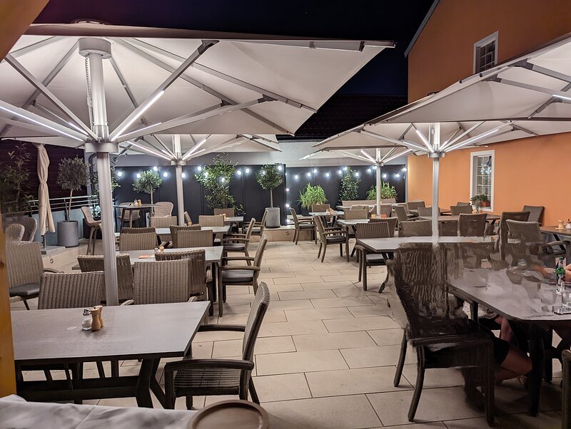 Au&szlig;enbereich eines Restaurants mit Tischen, St&uuml;hlen, gro&szlig;en Sonnenschirmen und Beleuchtung bei Nacht.