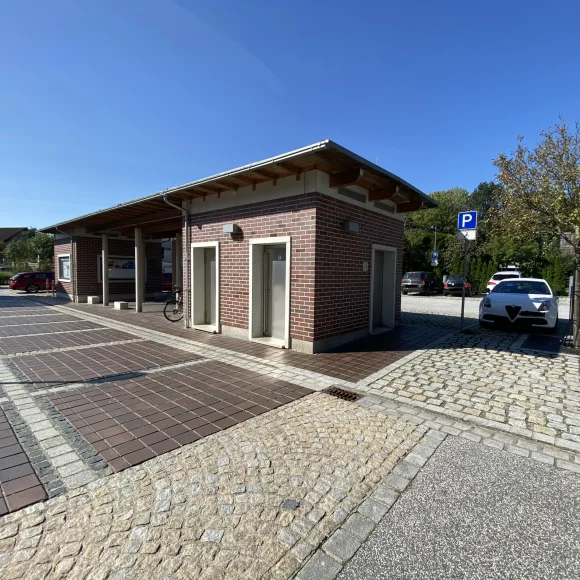 Gebäude mit drei Türen auf gepflastertem Platz bei klarem Himmel und geparktem Auto neben Baum.