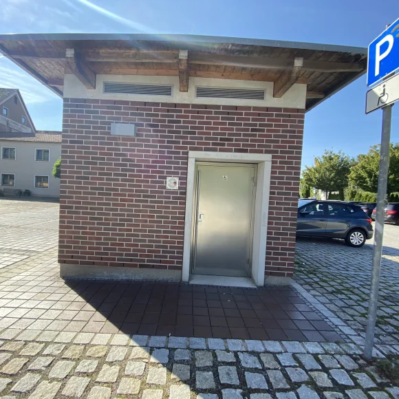 Kleines Gebäude mit Metalltür und Rollstuhlsymbol, daneben Parkplatz mit Behindertenparkplatzschild.