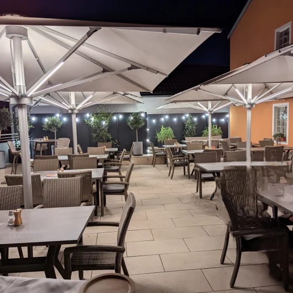 Außenbereich eines Restaurants mit Tischen, Stühlen, großen Sonnenschirmen und Beleuchtung bei Nacht.