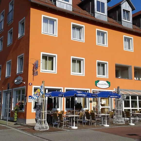 Oranger mehrstöckiger Gebäudekomplex mit Außengastronomie und blauen Sonnenschirmen an sonnigem Tag.