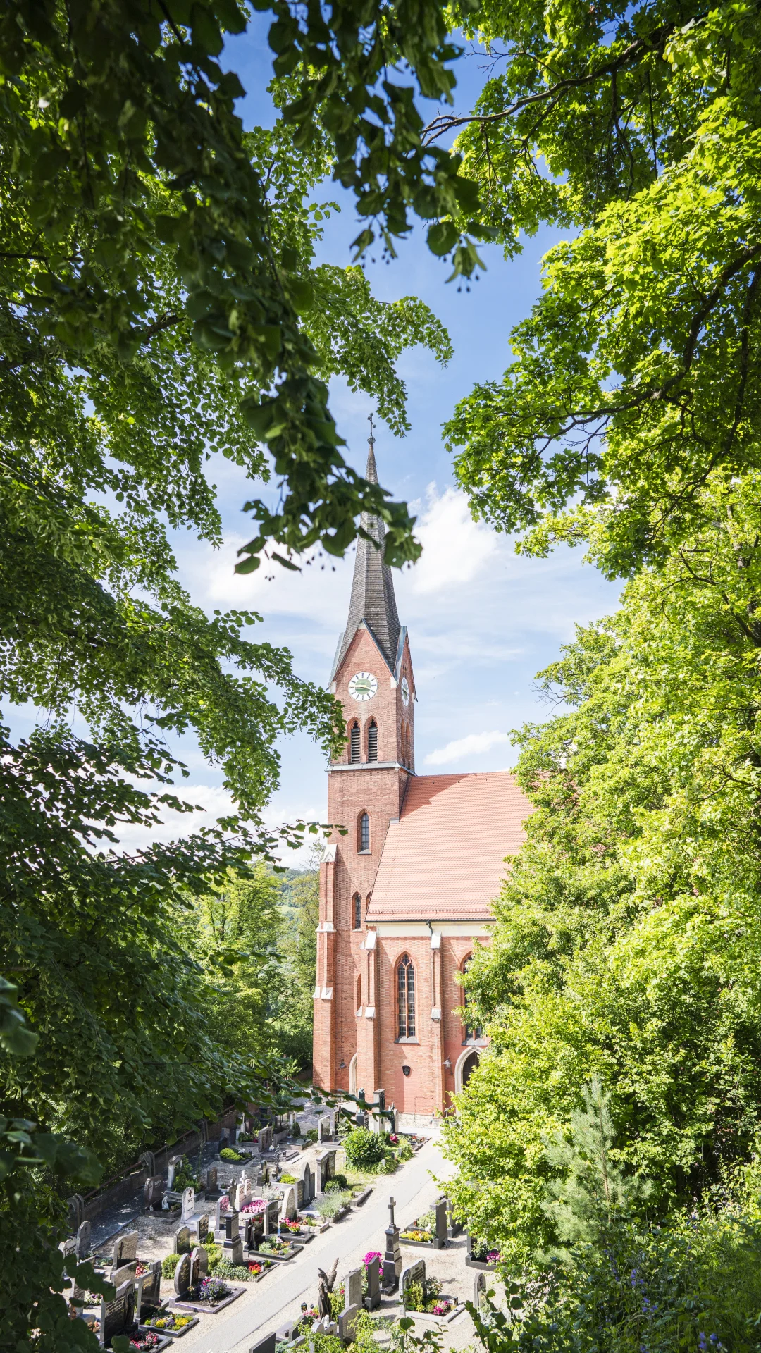 Rote Backsteinkirche mit Turm und Uhr, umgeben von Bäumen und einem Friedhof im Vordergrund.