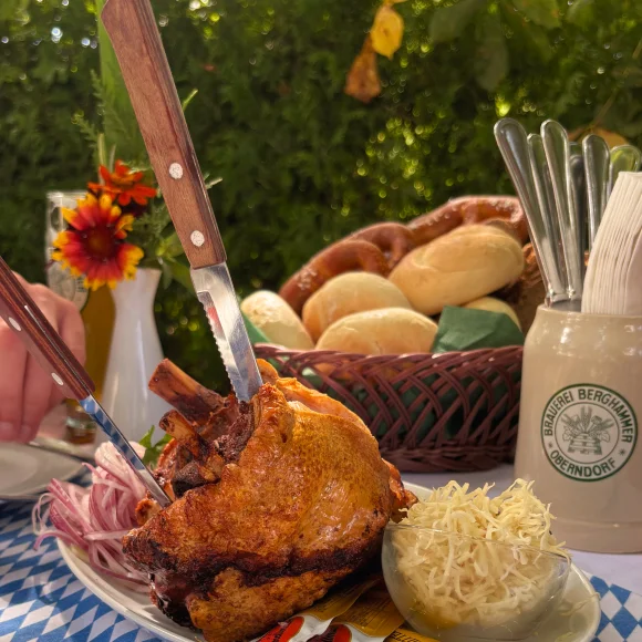 Teller mit Schweinshaxe, Krautsalat und Zwiebeln auf blau-weiß kariertem Tisch mit Brötchenkorb im Hintergrund.