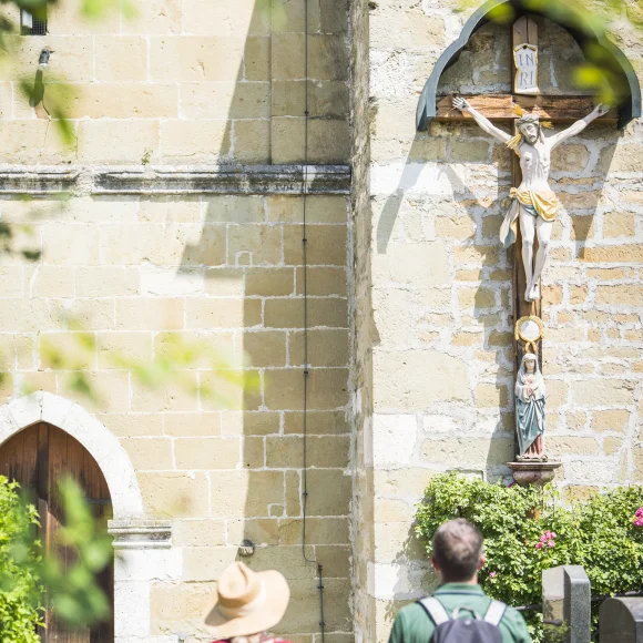 Zwei Wanderer gehen auf beiges Kirchengemäuer zu. An der Kirche hängt ein Jesuskreuz und eine braune alte Holztüre führt hinein. Es ist sonnig.