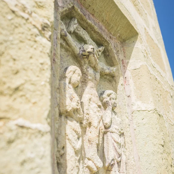Steinrelief an einer Wand, zeigt drei stehende Figuren unter einem Kreuz.