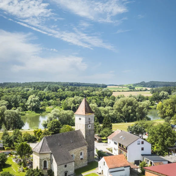 Das Luftbild zeigt die Kirche in Oberndorf aus erhöhter Perspektive. Die romanisch anmutende Kirche mit quadratischem Turm und rotem Zeltdach steht im Zentrum des Bildes. Umgeben ist sie von einem gepflegten Friedhof mit Grabsteinen und grünem Rasen. Direkt daneben befinden sich Wohnhäuser mit roten und grauen Dächern. Im Hintergrund fließt die Donau durch eine dicht bewaldete Uferlandschaft, dahinter erstrecken sich Felder, Wiesen und eine hügelige Waldkulisse. Der Himmel ist blau mit leichten Schleierwolken.