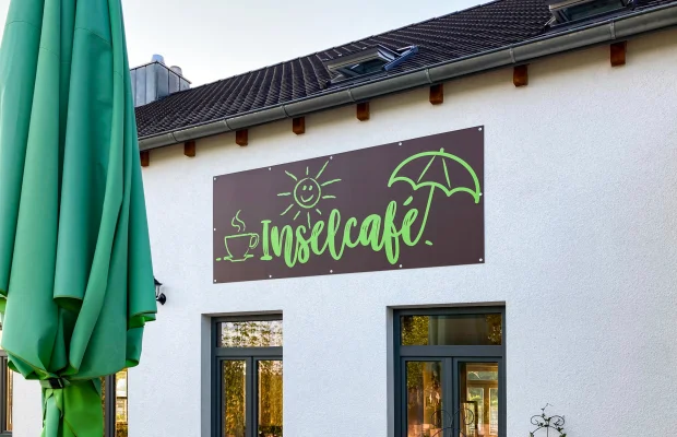 Außenansicht eines Gebäudes mit Schild „Inselcafé“ und geschlossenem grünem Sonnenschirm davor.