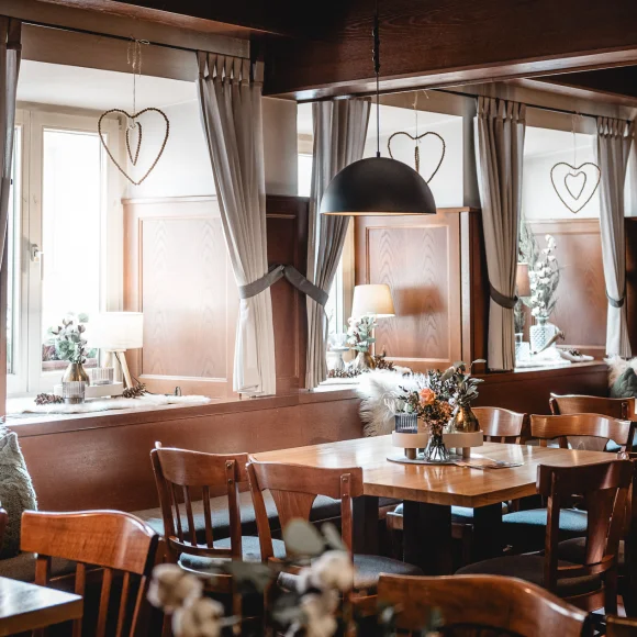 Gemütlicher Gastraum mit Holzmöbeln, Vorhängen, dekorativen Kissen und Blumen auf den Tischen.