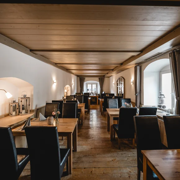 Restaurant mit Holztischen und schwarzen Stühlen, Holzboden und Decke, Wandleuchten, Fenster mit Vorhängen.