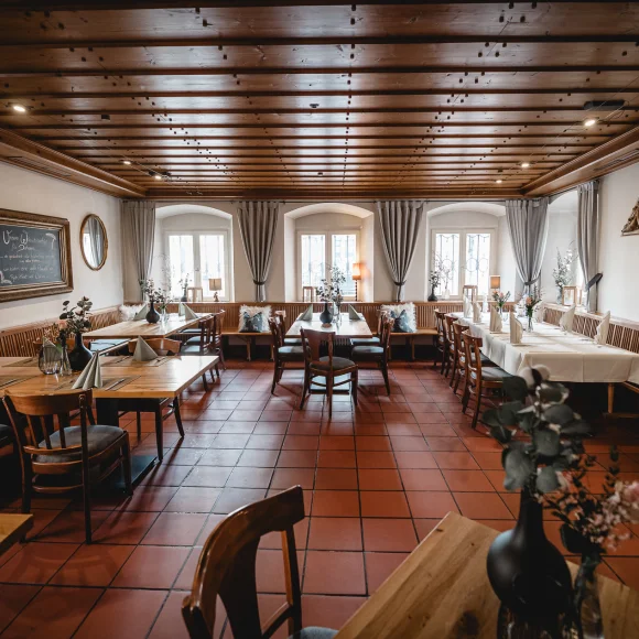 Ein leerer Speiseraum mit Holztischen, Stühlen, weißen Tischdecken und dekorativen Pflanzen auf den Tischen.